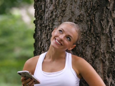 Mackenzie McKee leans against a tree and smiles while wearing a white tank top.