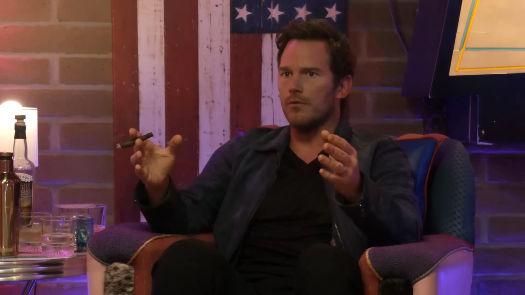 Chris Pratt gestures while holding a cigar and in front of a painted American flag.