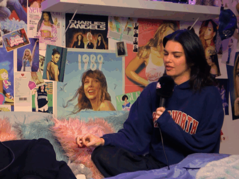 Kendall Jenner sits on a bed in front of a collage of posters of sensible music tastes and films, all from Owen Thiele.