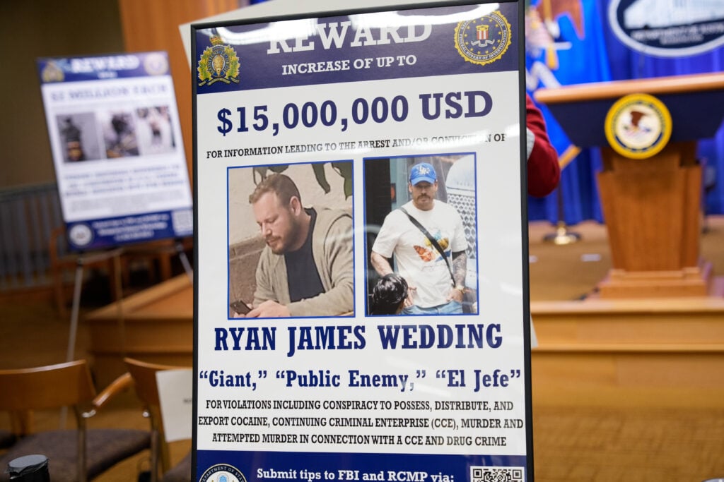A reward poster for the arrest of Ryan James Wedding is visible following a news conference announcing the indictment of a former Olympic snowboarder Ryan Wedding, charging him with murder and money laundering in connection to a drug trafficking organization at the Justice Department on November 19, 2025 in Washington, DC.